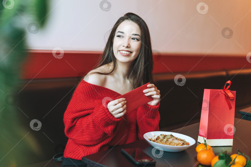 Скачать Красивая молодая азиатка в красной одежде ест азиатскую еду с красным конвертом в ресторане, празднуя китайский Новый год фотосток Ozero