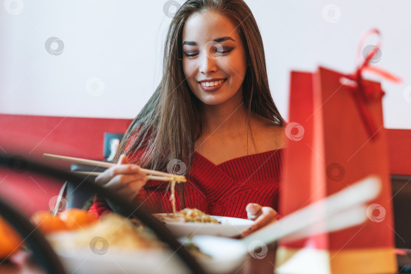 Скачать Красивая улыбающаяся молодая азиатка в красной одежде ест лапшу бамбуковыми палочками для еды в китайско вьетнамском ресторане фотосток Ozero