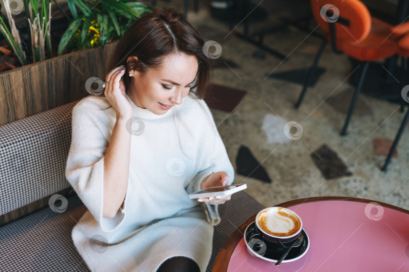 Скачать Привлекательная молодая брюнетка, улыбающаяся женщина в белом повседневном платье с чашкой кофе, пользующаяся мобильным телефоном в кафе фотосток Ozero