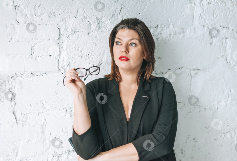Скачать Красивая улыбающаяся молодая женщина бизнес-леди в очках и черной куртке с мобильным телефоном на фоне белой стены фотосток Ozero