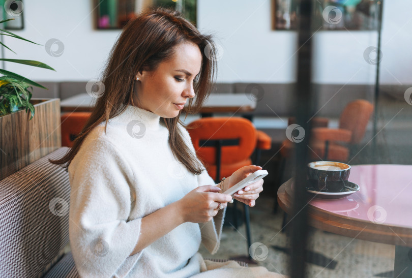 Скачать Привлекательная молодая брюнетка, улыбающаяся женщина в белом повседневном платье с чашкой кофе, пользующаяся мобильным телефоном в кафе фотосток Ozero