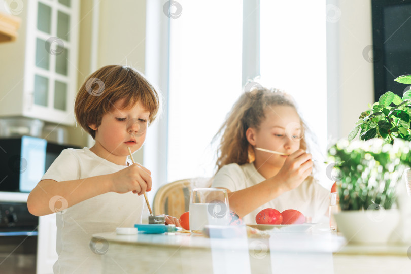 Скачать Два брата и сестра брат и сестра-малыш мальчик и девочка-подросток раскрашивают пасхальные яйца на кухне дома весенним солнечным днем фотосток Ozero