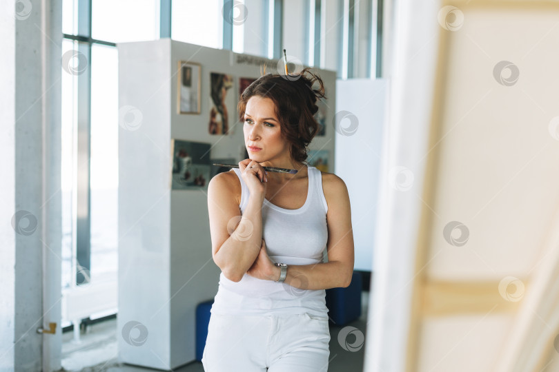 Скачать Размышляющая очаровательная молодая женщина-художник в белой одежде с кистями перед мольбертом в галерее современного искусства фотосток Ozero