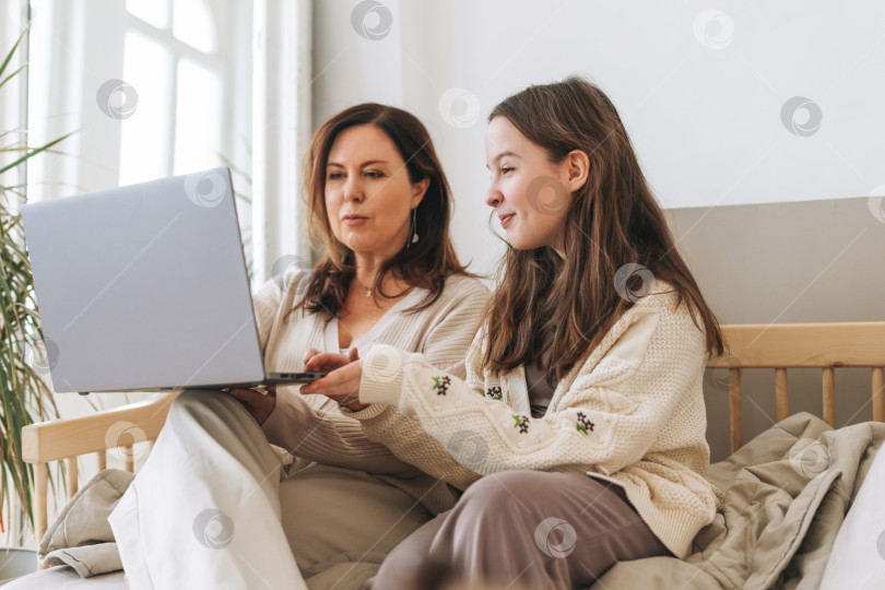 Скачать Мать, женщина средних лет, и дочь-подросток вместе пользуются ноутбуком в светлом интерьере фотосток Ozero