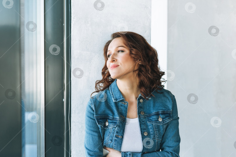 Скачать Портрет улыбающейся очаровательной молодой женщины в джинсовой куртке возле окна в современном здании фотосток Ozero