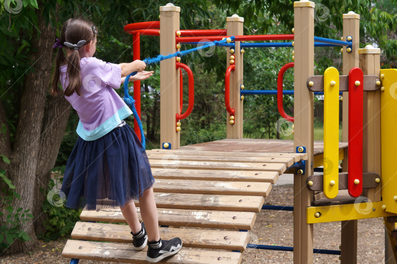 Скачать Саратов, Россия - 01 июня 2024 года. Девочка с хвостиками ловко забирается по канату на детской спортивной площадке фотосток Ozero