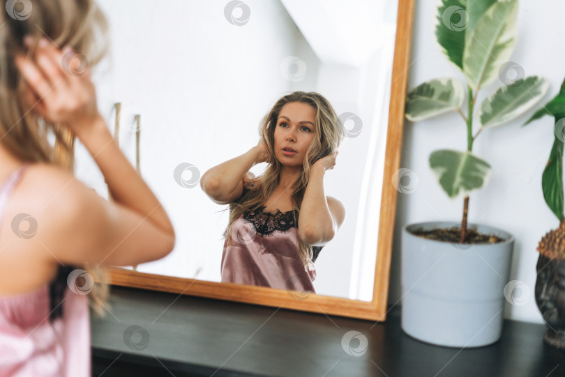 Скачать Светловолосая чувственная молодая женщина 35 лет с чистым свежим лицом и руками, с длинными волосами, в домашней одежде, смотрящая в зеркало на яркий интерьер фотосток Ozero