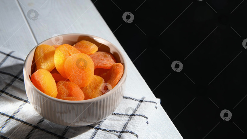 Скачать Женская рука берет курагу из миски, разложенной на клетчатой салфетке на белом деревянном столе, готовая отведать полезную и вкусную закуску с небольшим пространством для копирования фотосток Ozero