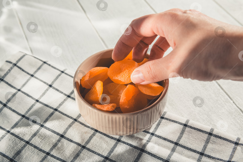 Скачать Женская рука тянется к миске с курагой, разложенной на клетчатой салфетке на белом деревянном столе, готовая насладиться полезной и вкусной закуской фотосток Ozero