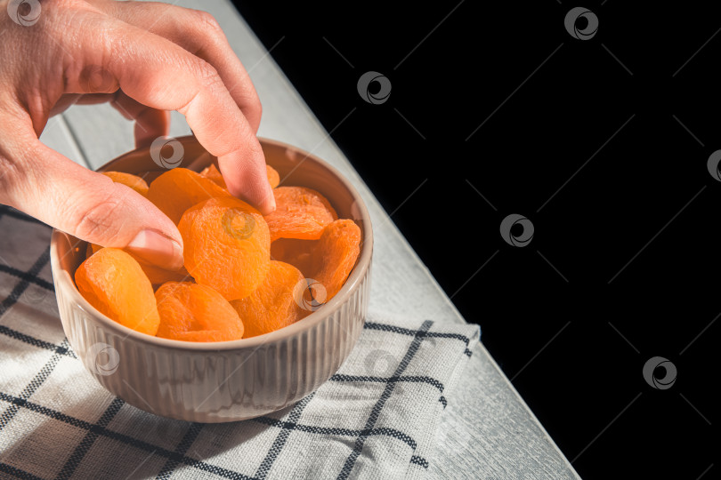 Скачать Рука выбирает курагу из керамической вазы, полной кураги, поставленной на клетчатую скатерть на белом деревянном столе на черном фоне. фотосток Ozero