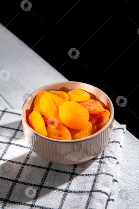 Скачать Аппетитная курага, разложенная в миске на клетчатой скатерти на белом деревянном столе, станет питательной закуской или ароматным ингредиентом для приготовления пищи. фотосток Ozero