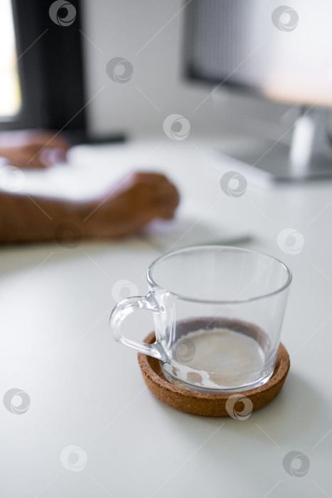 Скачать Кофейная чашка из прозрачного стекла на пробковой подставке рядом со взрослым африканским мужчиной, который печатает за письменным столом фотосток Ozero