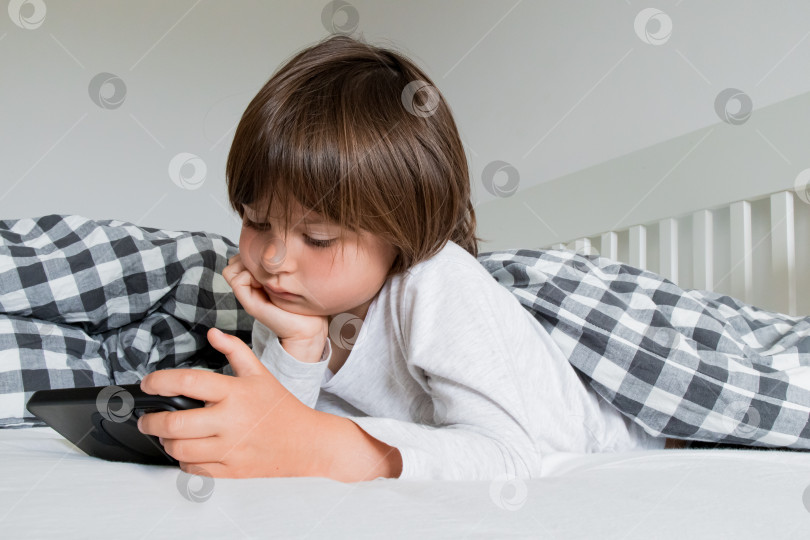 Скачать Молодой кавказский ребенок отдыхает в постели, глядя на планшет с клетчатым одеялом фотосток Ozero