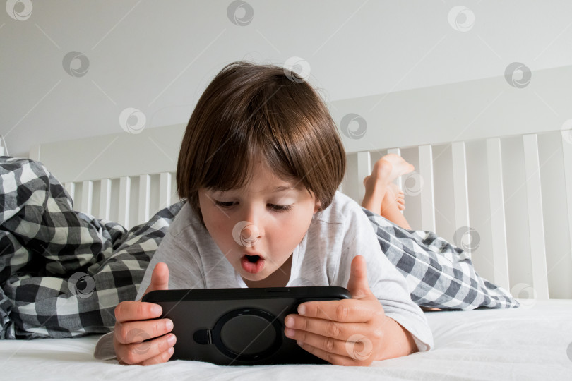 Скачать Молодой кавказский ребенок возбужденно играет на смартфоне в постели фотосток Ozero