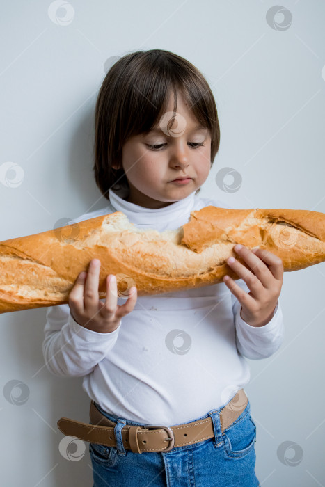 Скачать Молодой белый ребенок с длинными каштановыми волосами держит багет на фоне белой стены фотосток Ozero