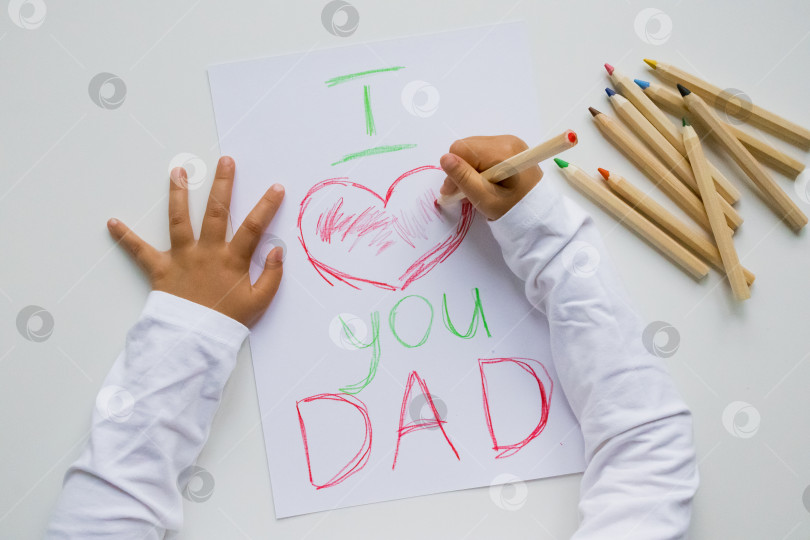 Скачать Ребенок рисует сердечное послание для отца цветными карандашами на белой бумаге. День отца фотосток Ozero
