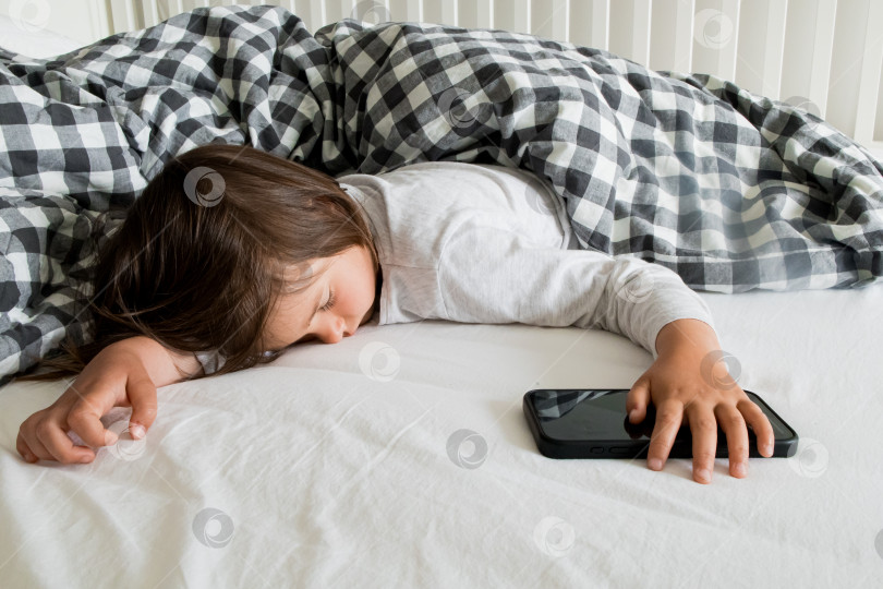 Скачать Молодой кавказский ребенок спит на кровати, держа смартфон под одеялом фотосток Ozero