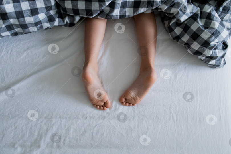 Скачать Детские ножки выглядывают из-под клетчатого одеяла на белой кровати фотосток Ozero