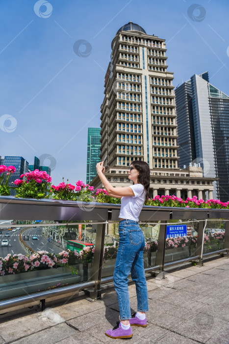 Скачать Молодая женщина фотографируется на пешеходном мосту в Пудуне, Шанхай, в окружении цветов и современных небоскребов в солнечный весенний день фотосток Ozero