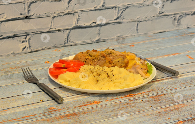 Скачать На деревянном столе тарелка с вкусным обедом, картофельное пюре, свинина, запеченная в сыре, и спелые помидоры, рядом столовые приборы. фотосток Ozero