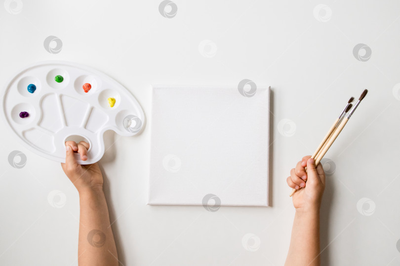 Скачать Детские руки держат палитру с красками и кисти рядом с чистым холстом фотосток Ozero