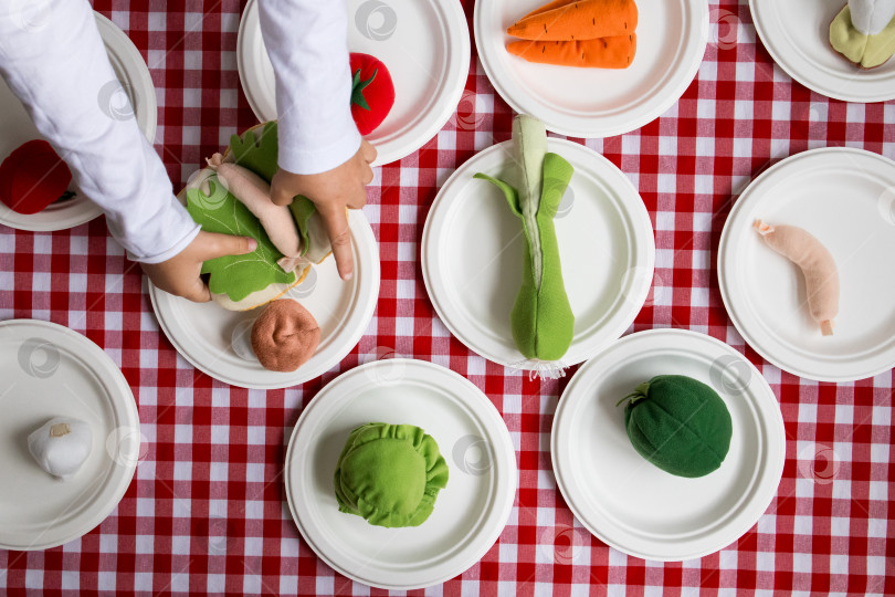Скачать Детские руки раскладывают тканевые овощи на столе для пикника с красно белой клетчатой скатертью фотосток Ozero