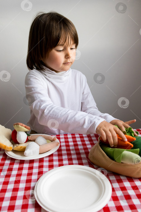 Скачать Кавказский ребенок играет с игрушечными фруктами и овощами на красной клетчатой скатерти фотосток Ozero