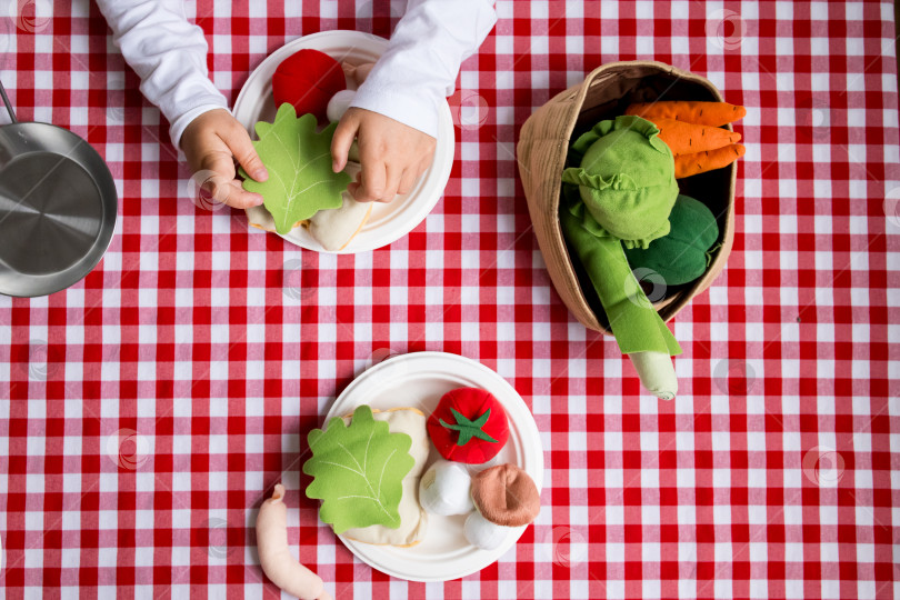 Скачать Ребенок играет с игрушечными овощами на красной клетчатой скатерти фотосток Ozero