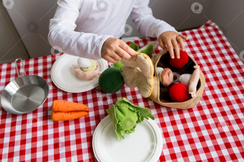 Скачать Ребенок играет с игрушечными овощами на красной клетчатой скатерти фотосток Ozero