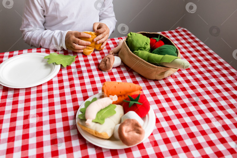 Скачать Ребенок играет с плюшевыми игрушечными продуктами на клетчатом столе на кухне фотосток Ozero