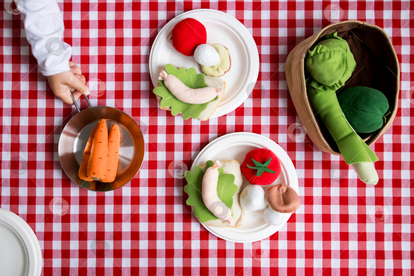 Скачать Ребенок играет с тканевыми овощами на красной клетчатой скатерти фотосток Ozero