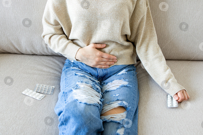 Скачать Молодая белая женщина с болью в животе сидит на диване с лекарствами в руках фотосток Ozero