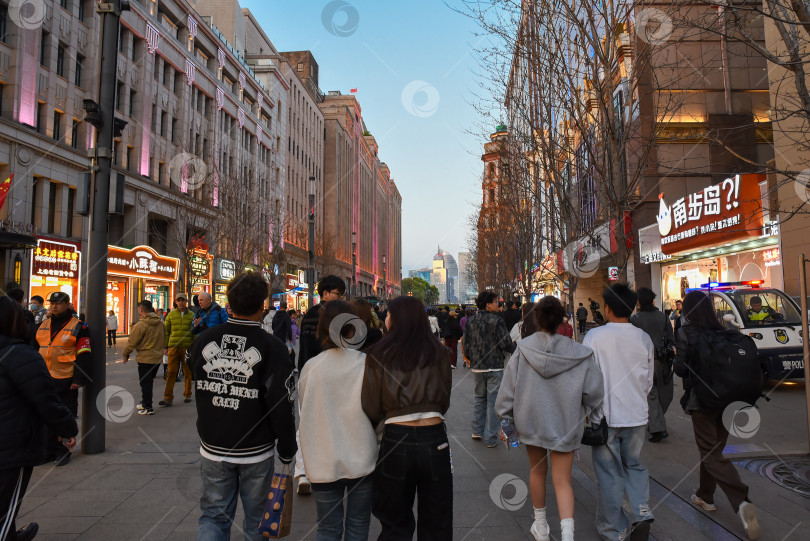 Скачать Шанхай, Китай - 1 апреля 2025 года: Молодежь и гости города прогуливаются и делают покупки по яркой Нанкинской улице со светящимися вывесками, продуктовыми киосками и неоновыми огнями в сумерках. фотосток Ozero