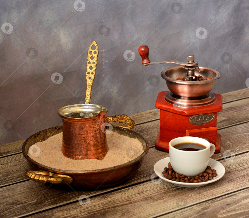 Скачать Чашка черного кофе в зернах, ручная кофемолка и медная джезва в поддоне с песком на деревянном столе. фотосток Ozero