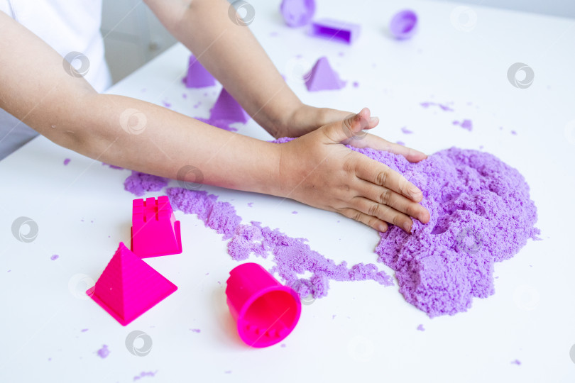 Скачать Ребенок играет с фиолетовым кинетическим песком и лепными фигурами на белом столе фотосток Ozero