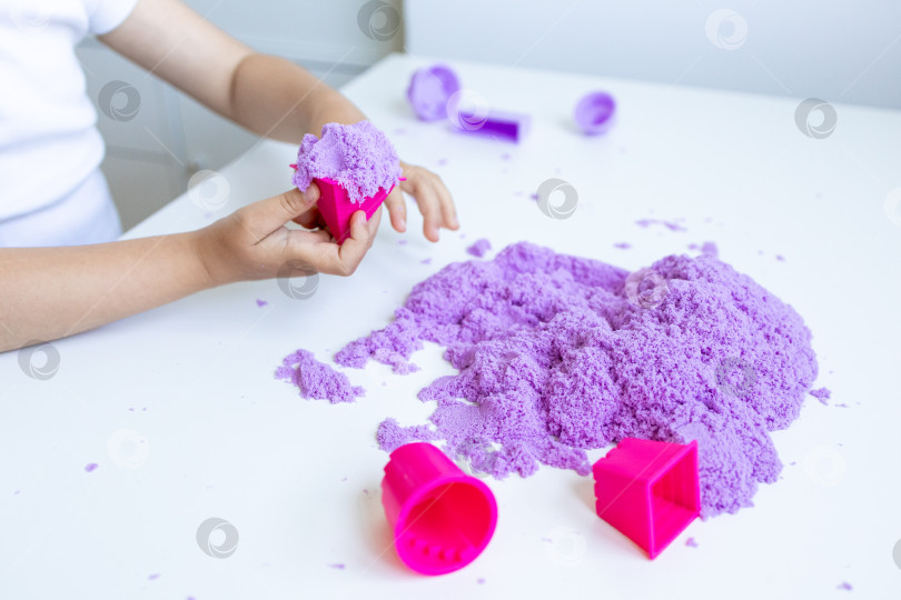 Скачать Ребенок играет с фиолетовым кинетическим песком и розовыми формочками на белом столе фотосток Ozero