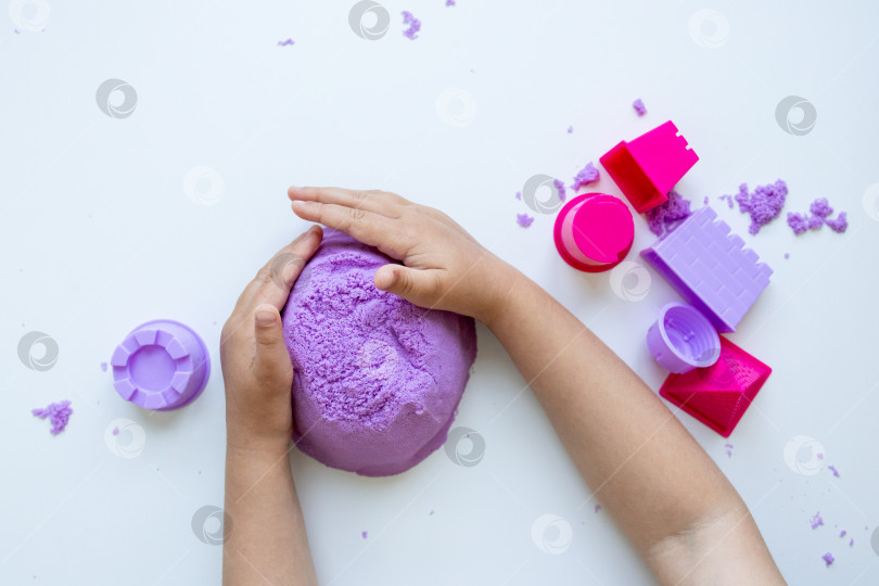 Скачать Детские руки лепят фиолетовый кинетический песок из разноцветных формочек на белом фоне фотосток Ozero