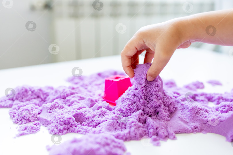 Скачать Детская рука играет с фиолетовым кинетическим песком и розовой плесенью на столе фотосток Ozero