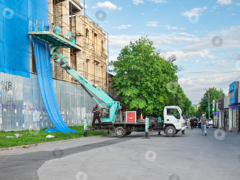 Скачать Гидроподъемник (краново-механическая установка) на базе грузовика Isuzu Elf для строительного объекта. Рабочие драпируют здание фотосток Ozero