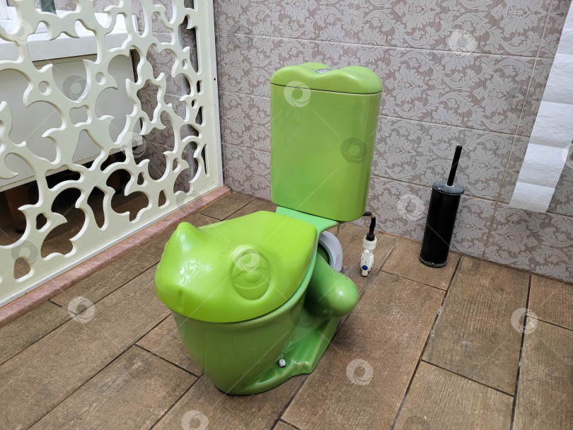Скачать В ванной комнате установлен зеленый детский унитаз в форме лягушки на фоне бежевой плитки. фотосток Ozero
