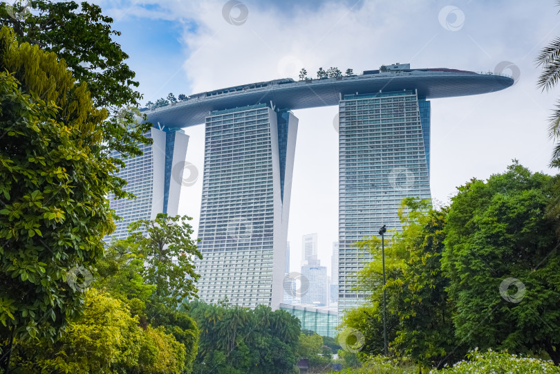 Скачать Сингапур, Сингапур – 4 апреля 2025 года: Вид на знаменитый отель Marina Bay Sands, окруженный тропической зеленью, с его небоскребом Sky park, раскинувшимся на трех башнях. фотосток Ozero