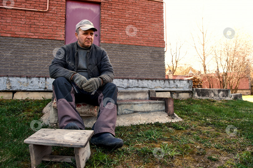 Скачать Взрослый мужчина пенсионного возраста сидит на ступеньке и смотрит в сторону. Фермер. фотосток Ozero