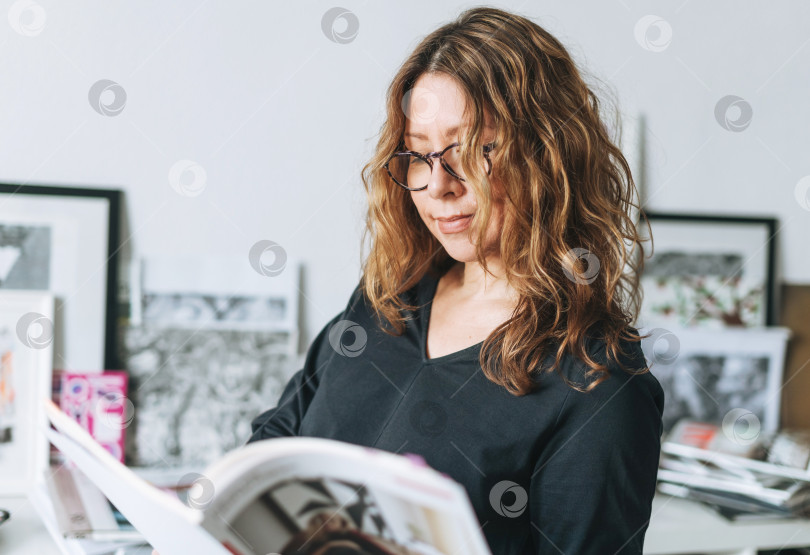 Скачать Очаровательная улыбающаяся взрослая женщина средних лет с вьющимися волосами архитектор дизайнер с журнальным каталогом в руках в современном офисе фотосток Ozero