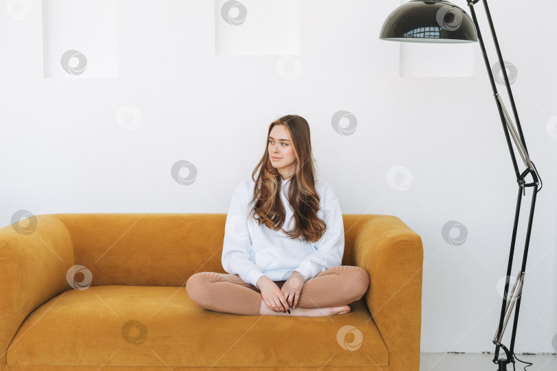 Скачать Молодая женщина подросток в белой толстовке с капюшоном красивая девушка сидит на желтом диване в современном интерьере фотосток Ozero