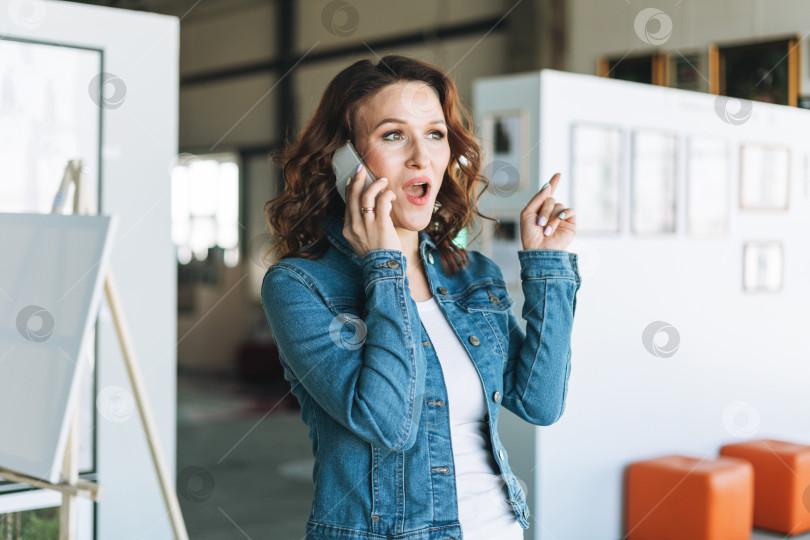 Скачать Очаровательная эмоциональная молодая женщина бизнесвумен дизайнер художник в джинсовой куртке, пользующаяся мобильным телефоном в галерее современного искусства фотосток Ozero