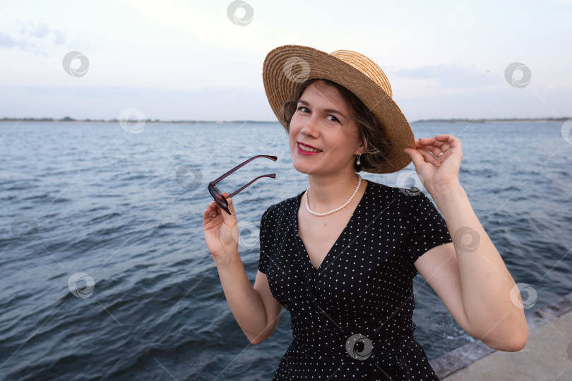 Скачать Женщина улыбается на берегу моря, держа в руках солнечные очки и соломенную шляпу. фотосток Ozero