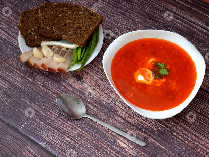 Скачать Борщ, вкусный и питательный суп, рядом с ним тарелка с ржаным хлебом, копченым салом, чесноком и зеленым луком и столовая ложка на деревянном столе. фотосток Ozero