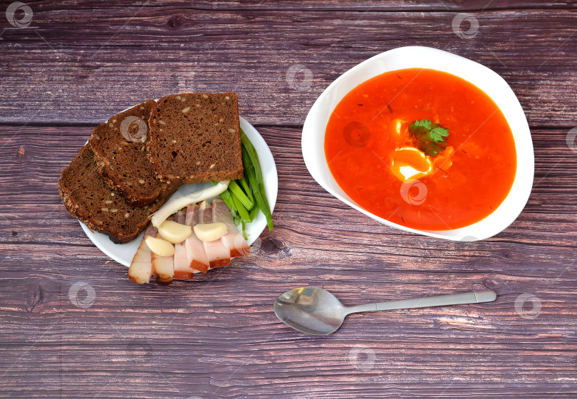 Скачать На деревянном столе - глубокая тарелка вкуснейшего славянского борща со сметаной и петрушкой, рядом - ложки и тарелка с ржаным хлебом, салом, чесноком и зеленым луком. фотосток Ozero