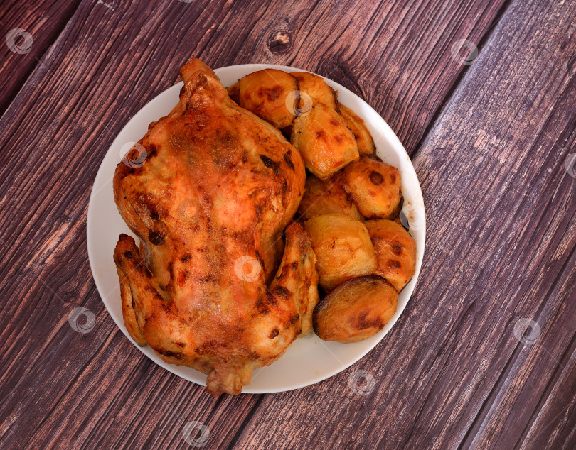 Скачать Запеченная целиком домашняя курица с молодым картофелем на большом круглом блюде на деревянном столе. фотосток Ozero