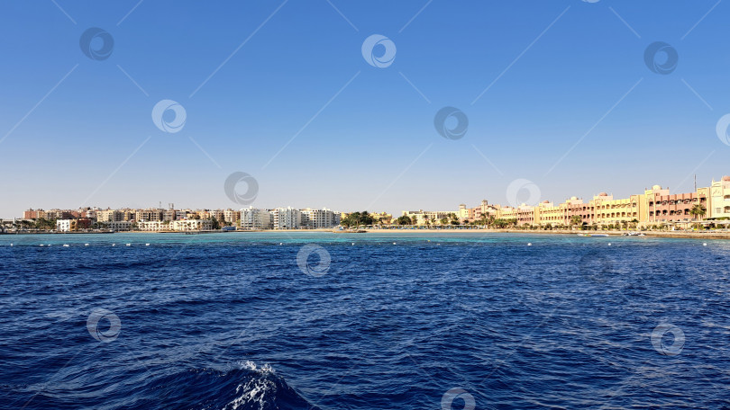 Скачать Прибрежный вид на оживленную береговую линию с чистыми голубыми водами и современными зданиями под ясным небом, создающими безмятежную пляжную атмосферу. Красное море. Хургада. Египет фотосток Ozero